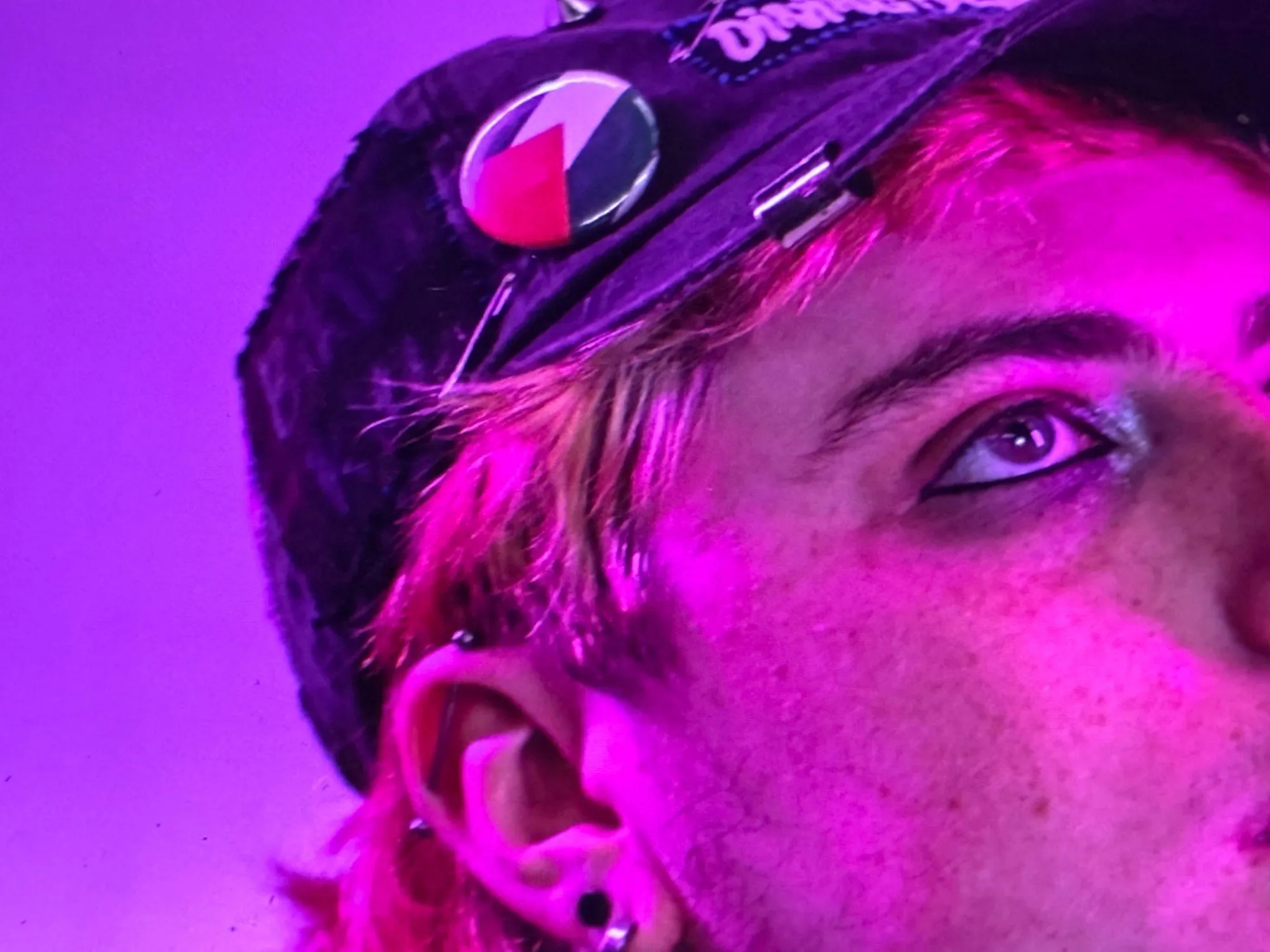 Close-up of a person in a cap with a pin, lit in magenta light, looking upward