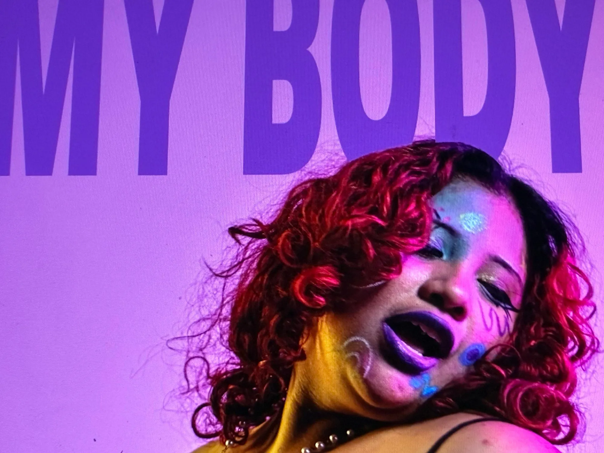 Person with red curly hair and glittery face paint beneath large purple letters reading "MY BODY"