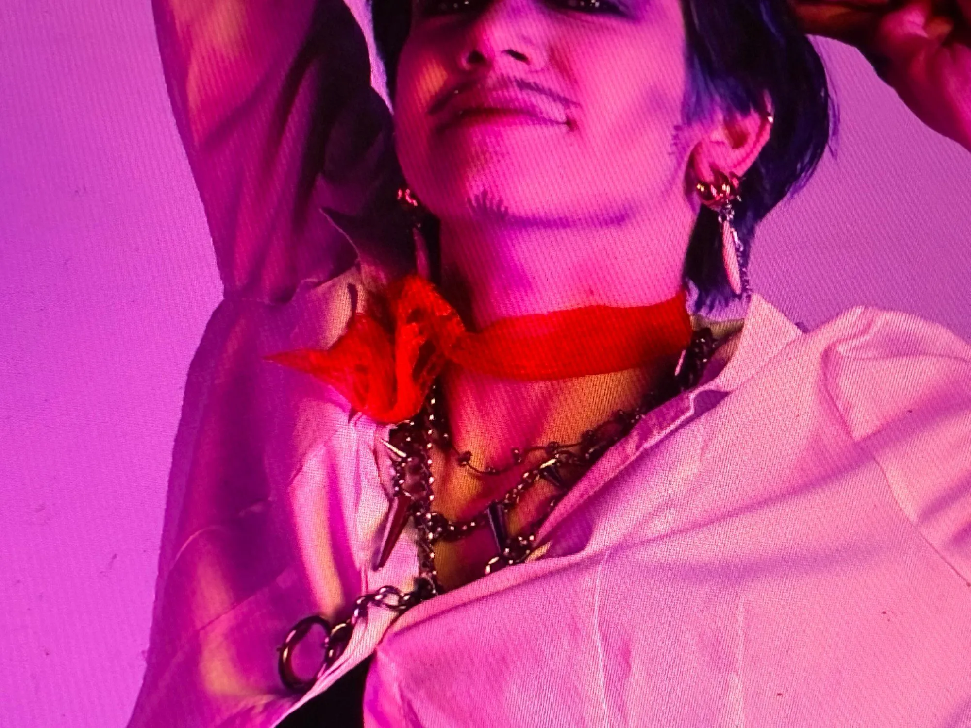 Person with their arm raised, wearing a red scarf, layered chains, and a white button-up shirt, lit in pink