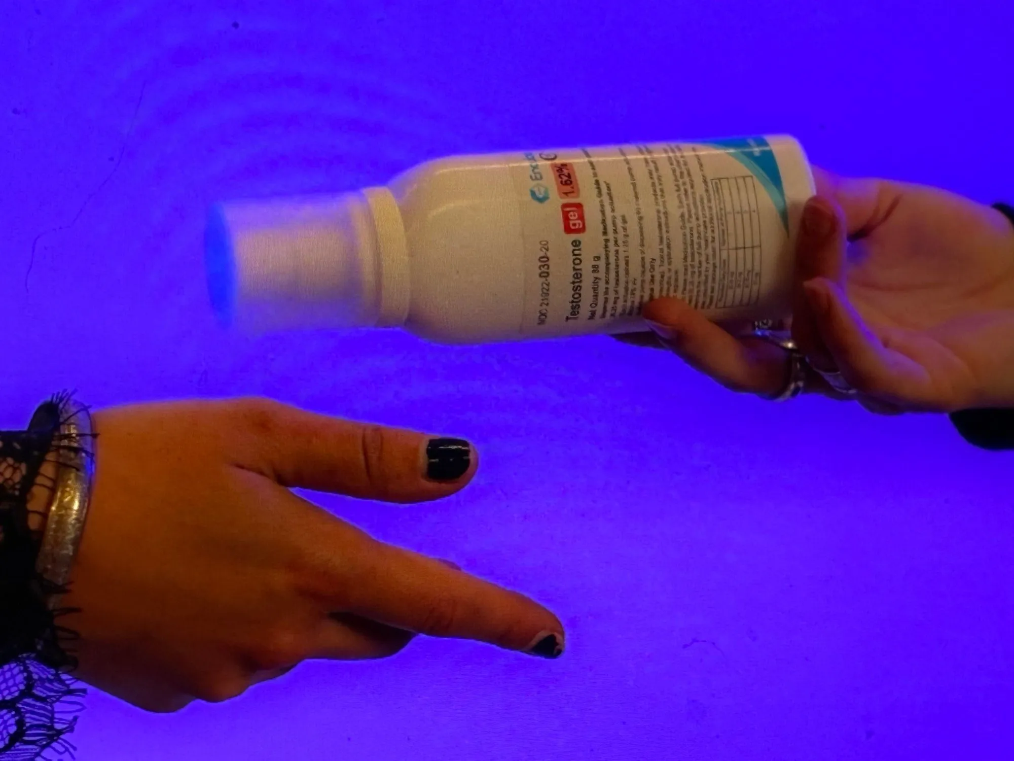 One hand holding a bottle of testosterone toward another hand pointing at it, under blue light