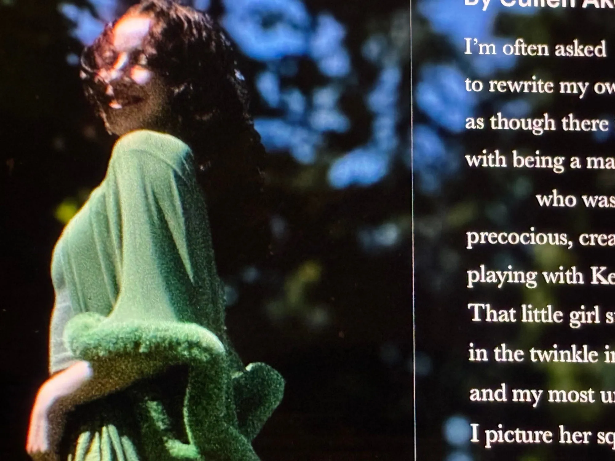 Person in a green dress with a fuzzy green wrap smiling in a garden, beside the start of an essay